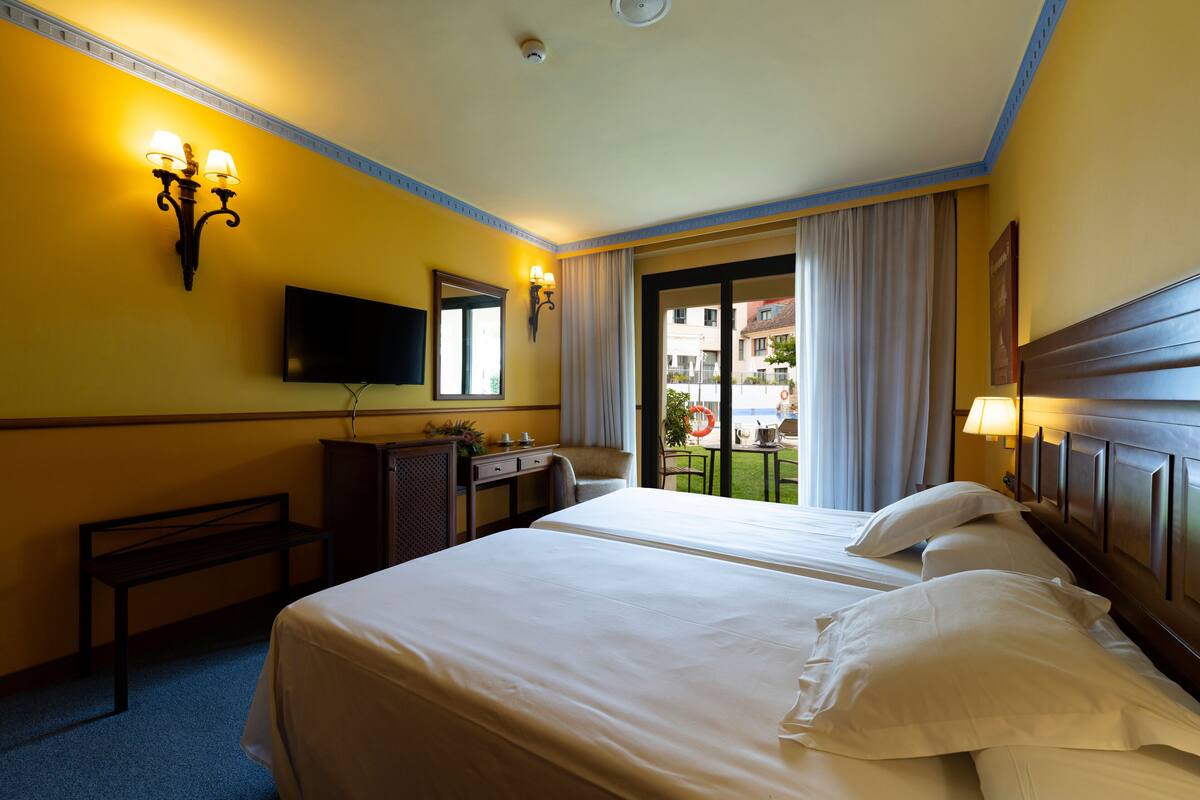 A hotel room with two beds, a desk, a chair, a television, and a balcony with a view of buildings.