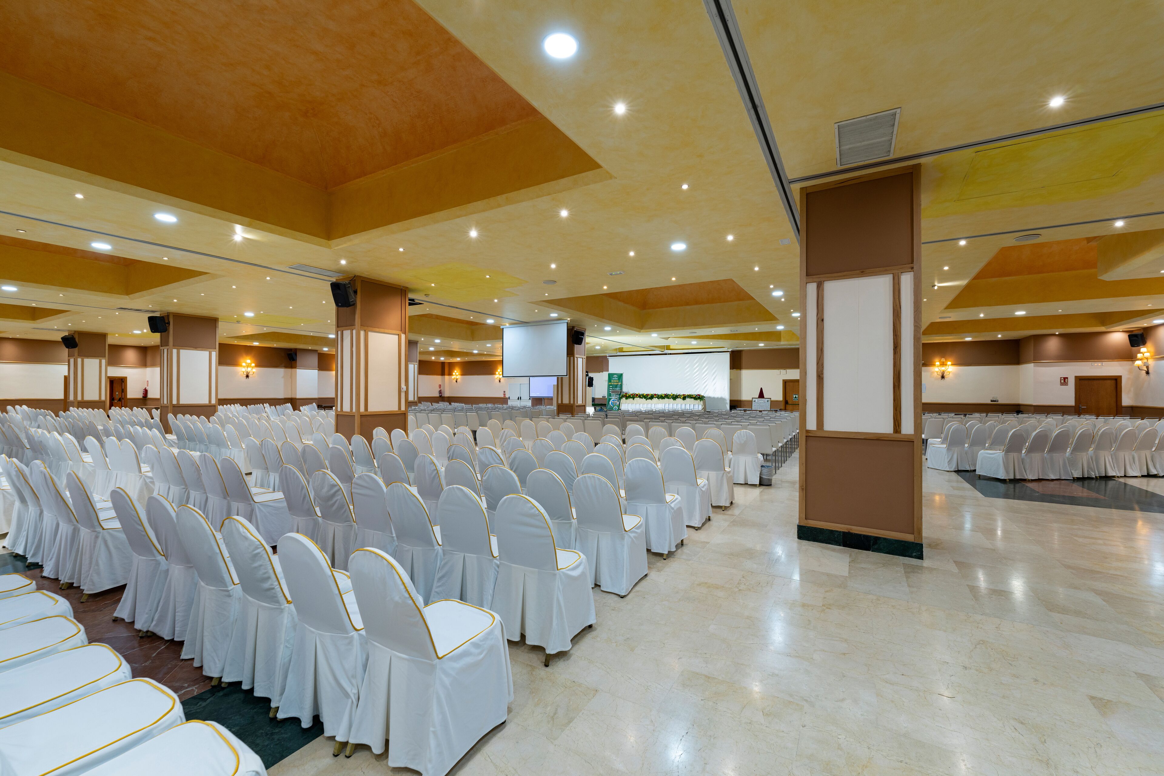 A large hall with rows of white chairs and tables, a stage with a screen, and a ceiling with recessed lighting.