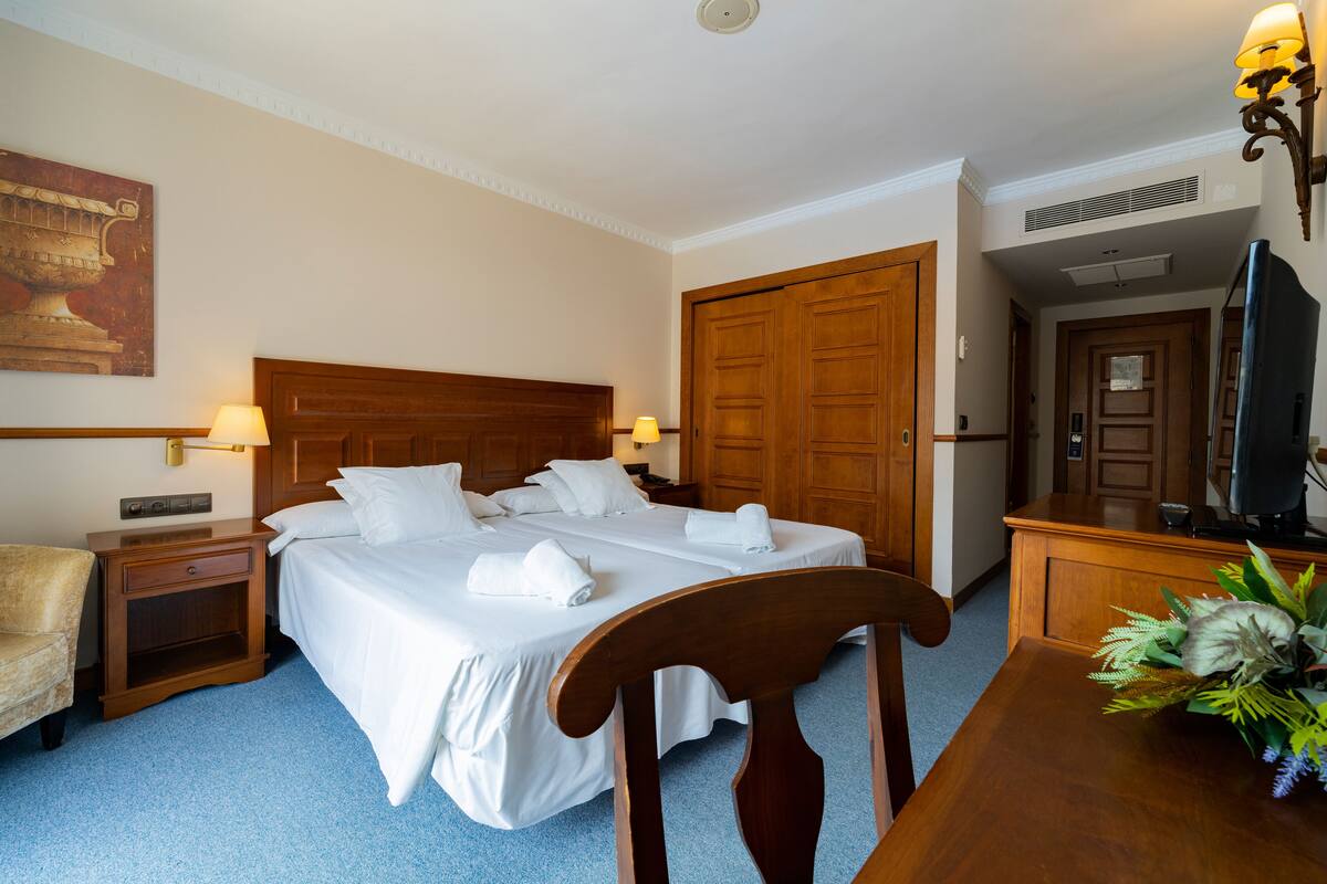 A hotel room with a large bed, wooden headboard, two bedside tables with lamps, a wooden desk, and a television.