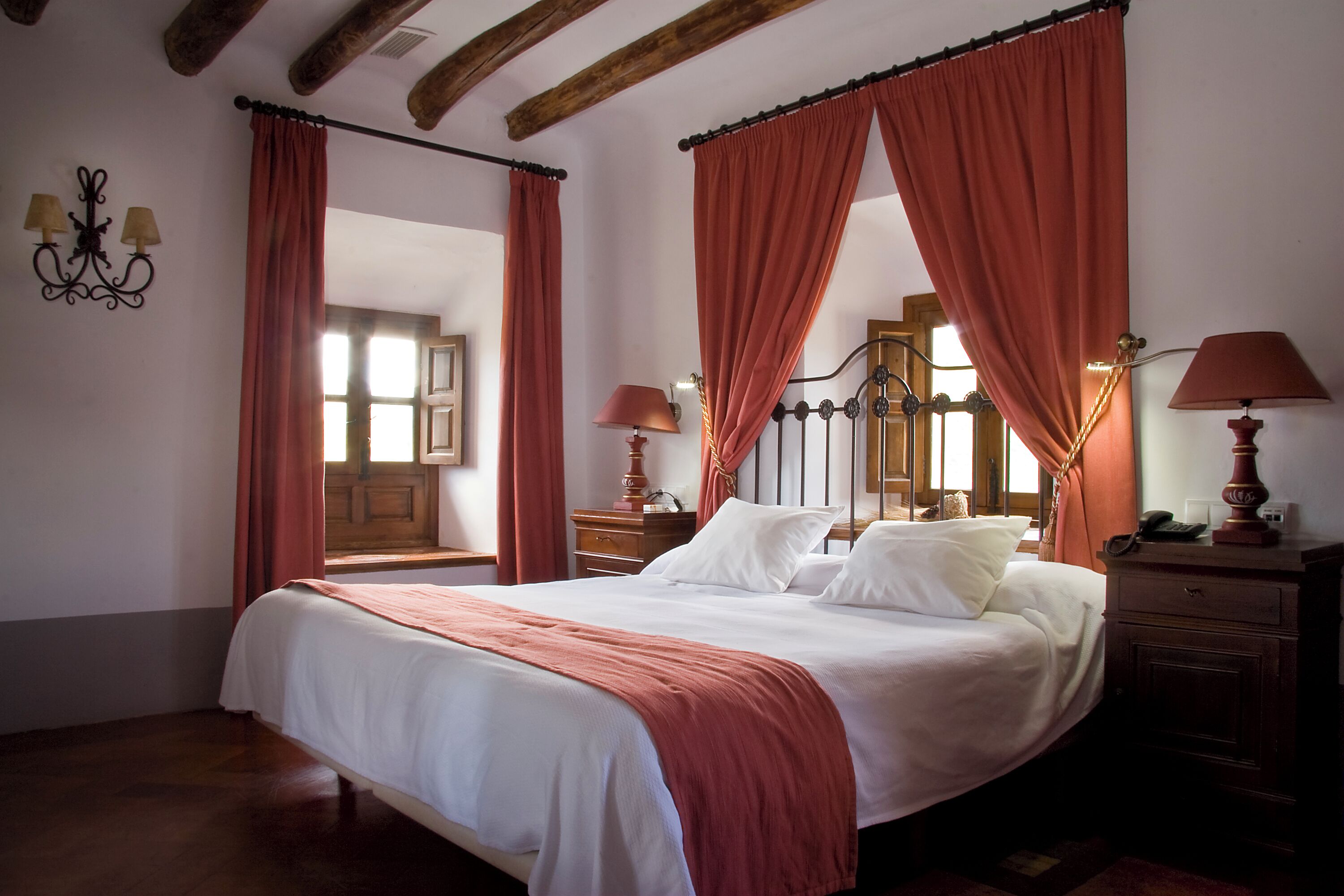 A bedroom with a large bed, wooden headboard, and red curtains. There are two bedside tables with lamps and a wooden nightstand.
