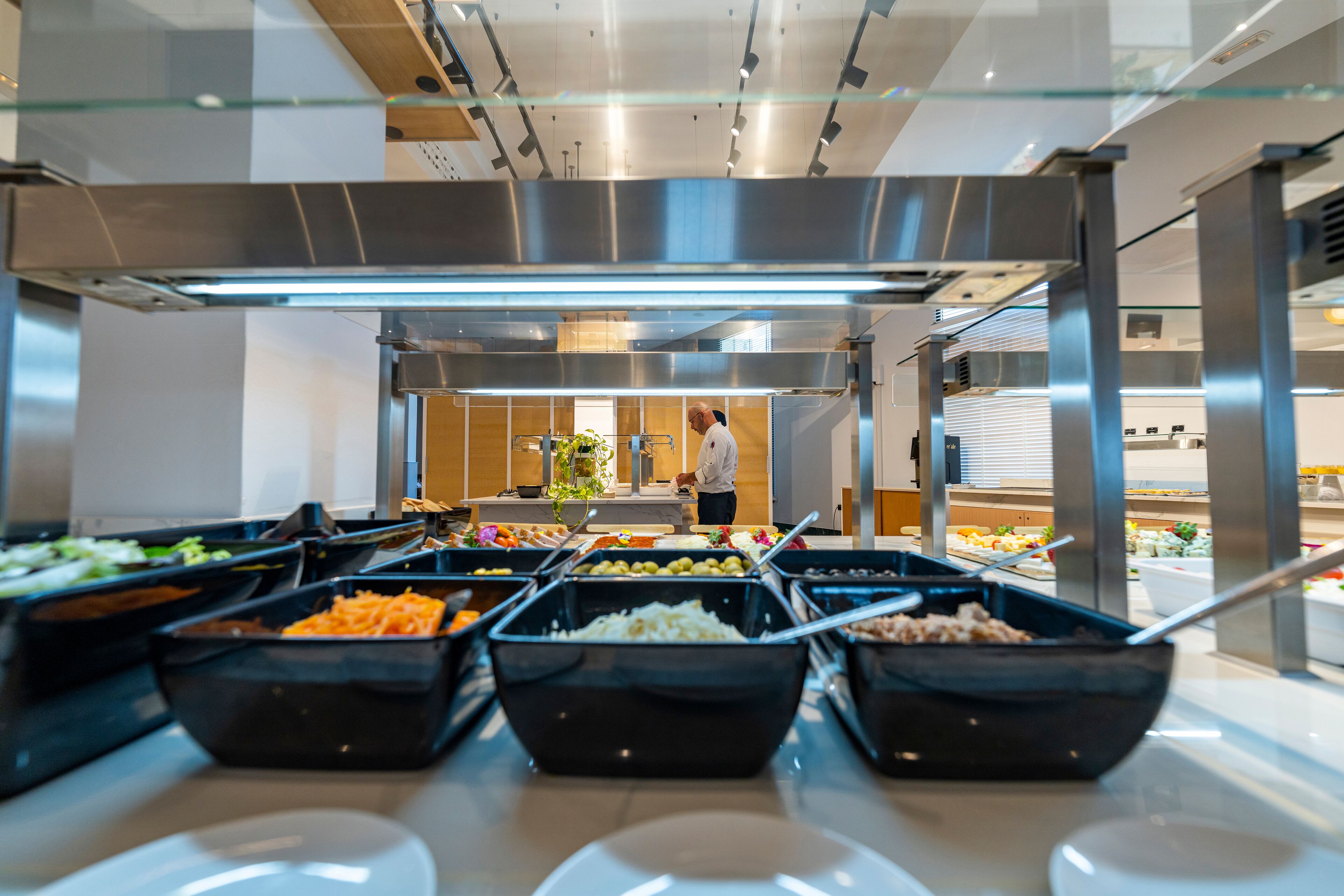 A buffet with various food options, a person serving, and a modern kitchen in the background.