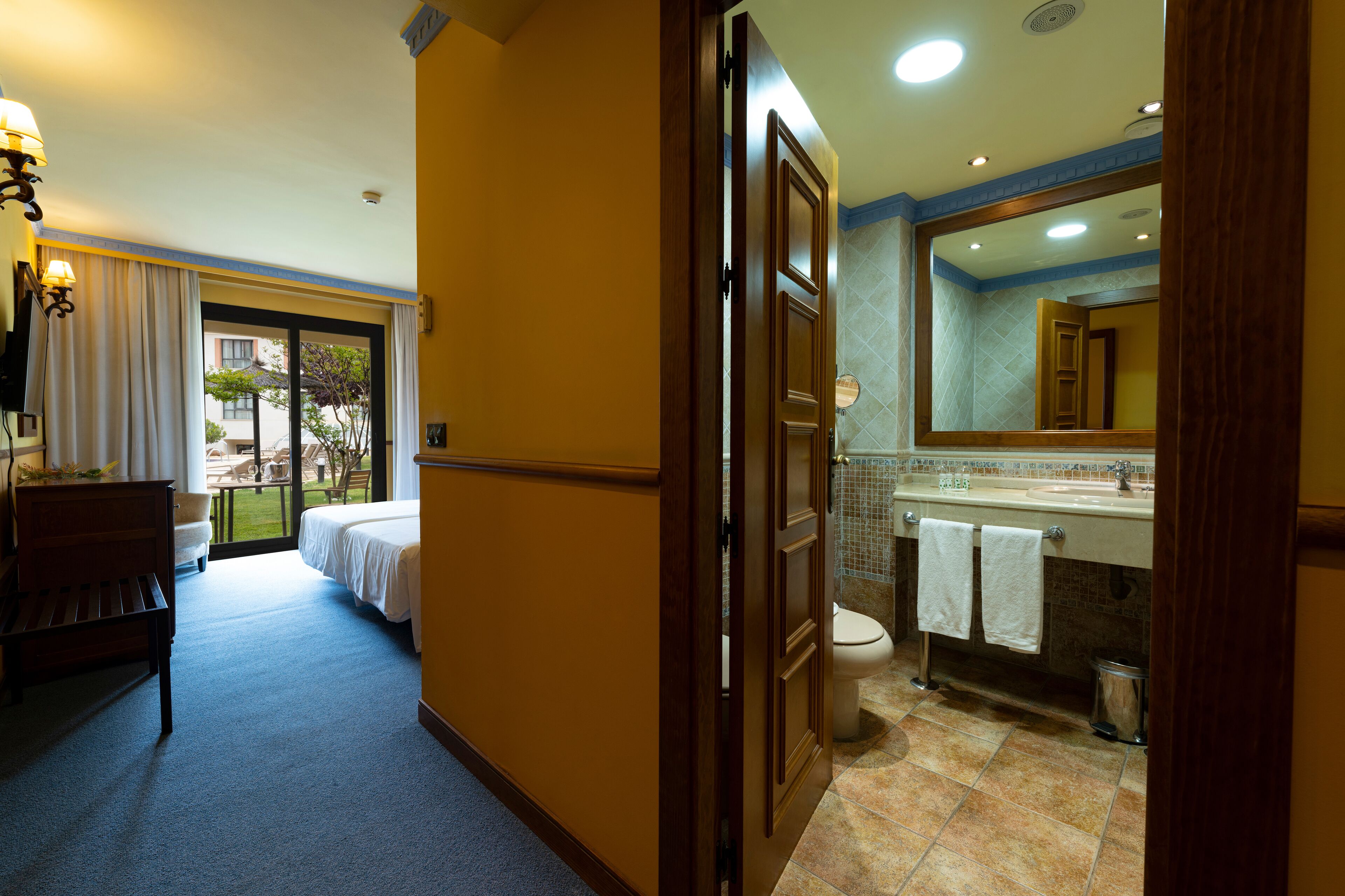 Habitación de hotel con cama, banco, escritorio, ventana con cortinas y baño con lavabo, espejo y toallero.