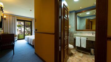 Habitación de hotel con cama, banco, escritorio, ventana con cortinas y baño con lavabo, espejo y toallero.