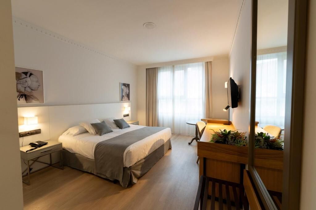 A hotel room with a bed, bedside table, lamp, TV, and a small desk with a chair.