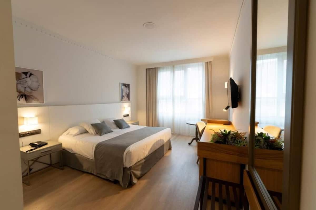 A hotel room with a bed, bedside table, lamp, TV, and a small desk with a chair.