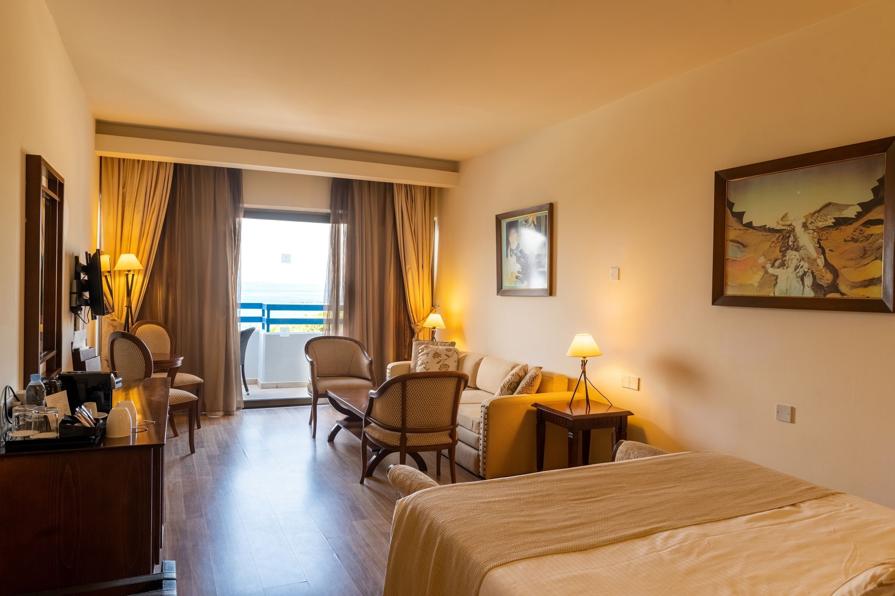 A hotel room with a bed, a desk, a chair, a sofa, and a view of the sea through large windows.