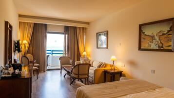 A hotel room with a bed, a desk, a chair, a sofa, and a view of the sea through large windows.