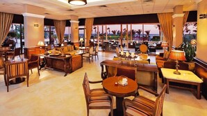 A spacious hotel lobby with multiple seating areas, including a central coffee table, sofas, and chairs. Large windows offer views of palm trees and the ocean. - Elias Beach Hotel (Limassol)