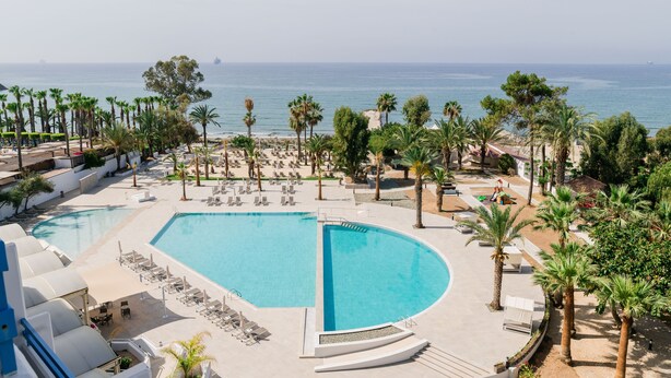 A large, curved swimming pool with lounge chairs and palm trees, overlooking a beach and ocean. - Elias Beach Hotel (Limassol)