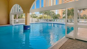 Una piscina interna con pareti in vetro trasparente, pavimento piastrellato e vista su alti edifici e palme circostanti.