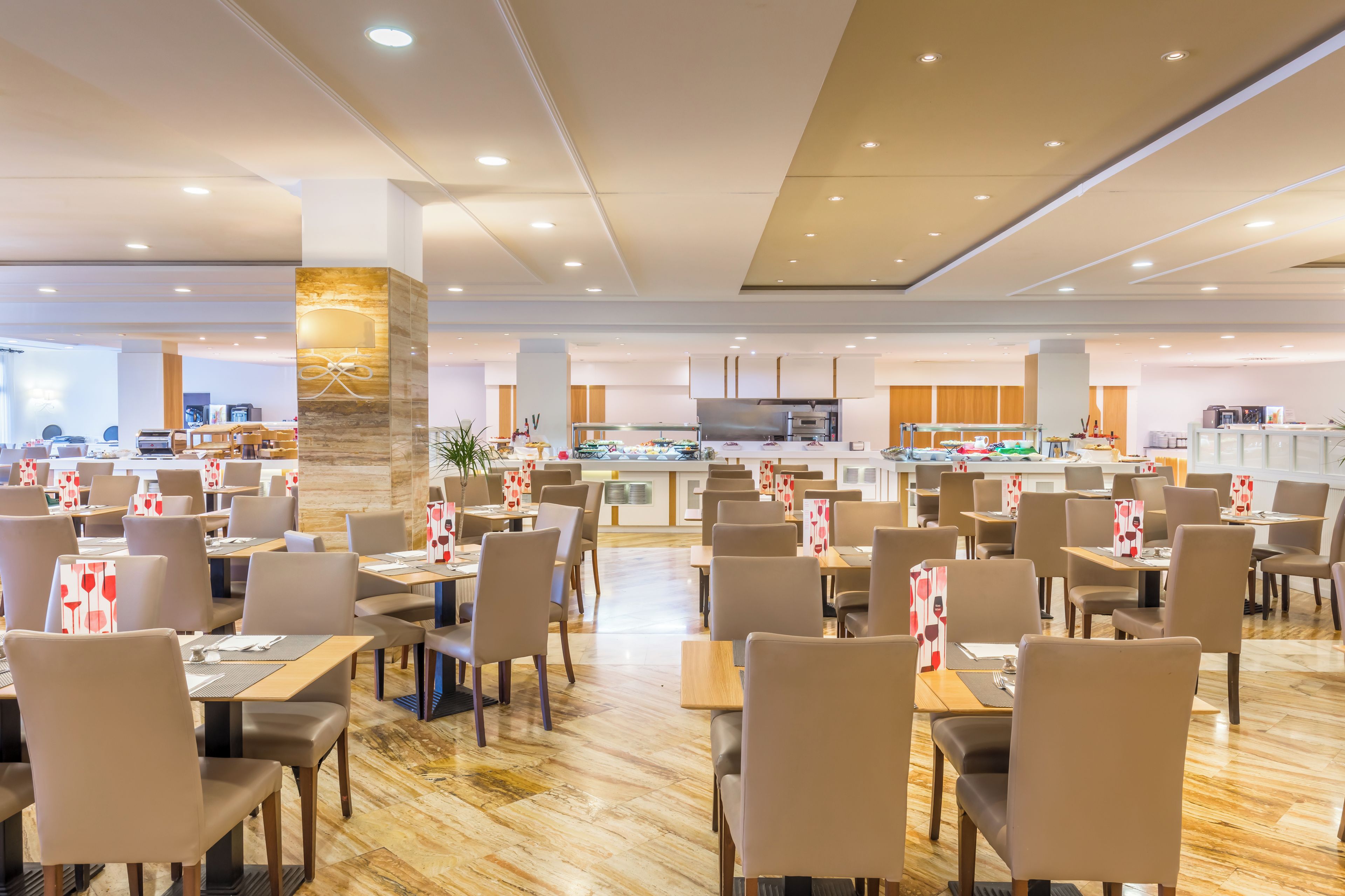 A spacious dining area with multiple tables and chairs, a central buffet, and a kitchen area in the background.