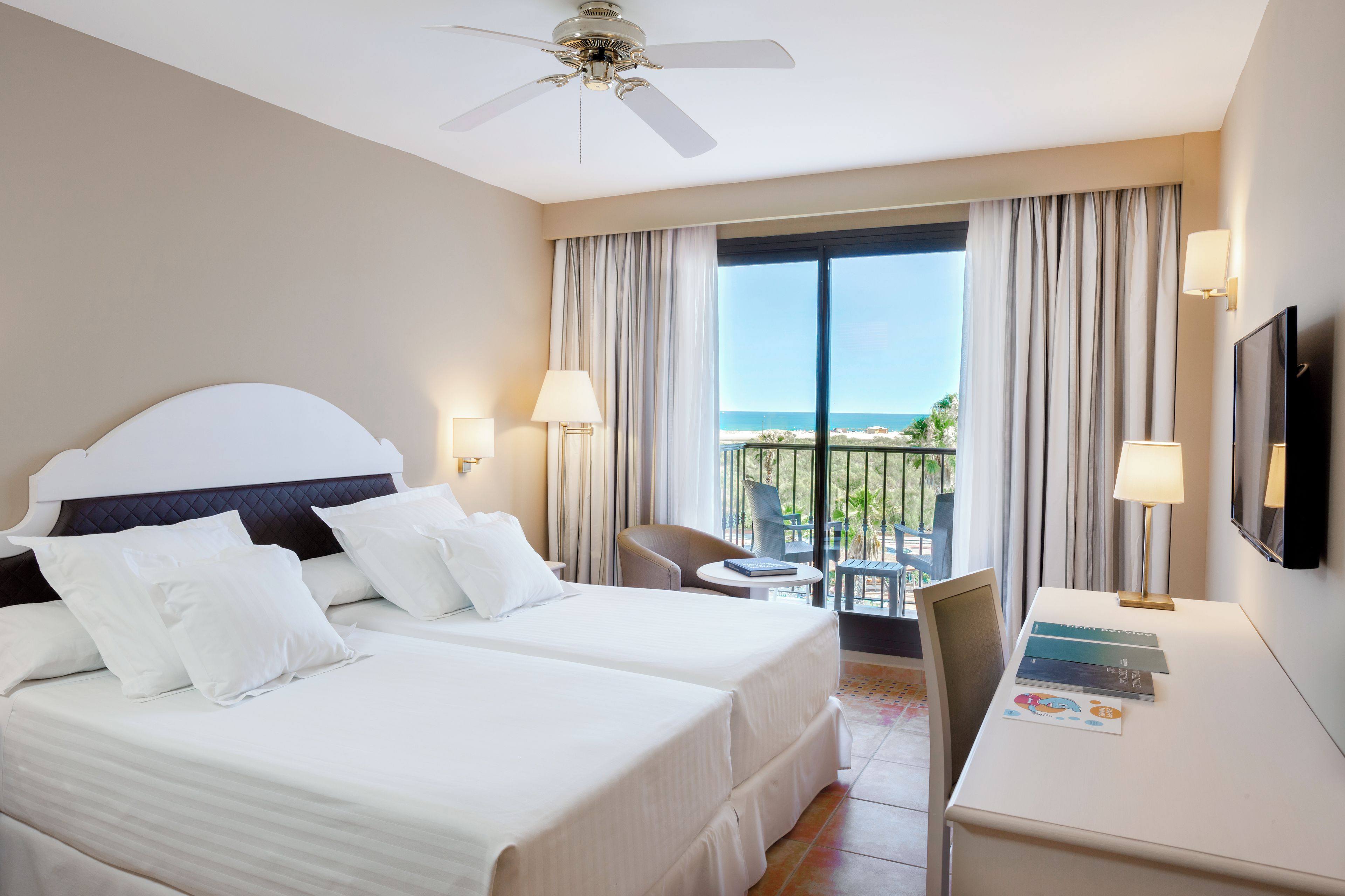 A hotel room with a bed, two bedside tables, a desk, a chair, a TV, and a balcony with a view of the sea.