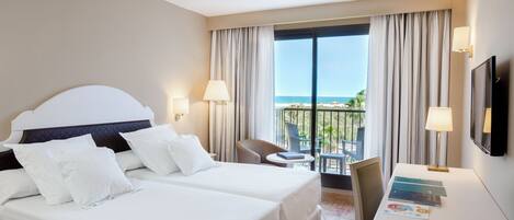 A hotel room with a bed, two bedside tables, a desk, a chair, a TV, and a balcony with a view of the sea.