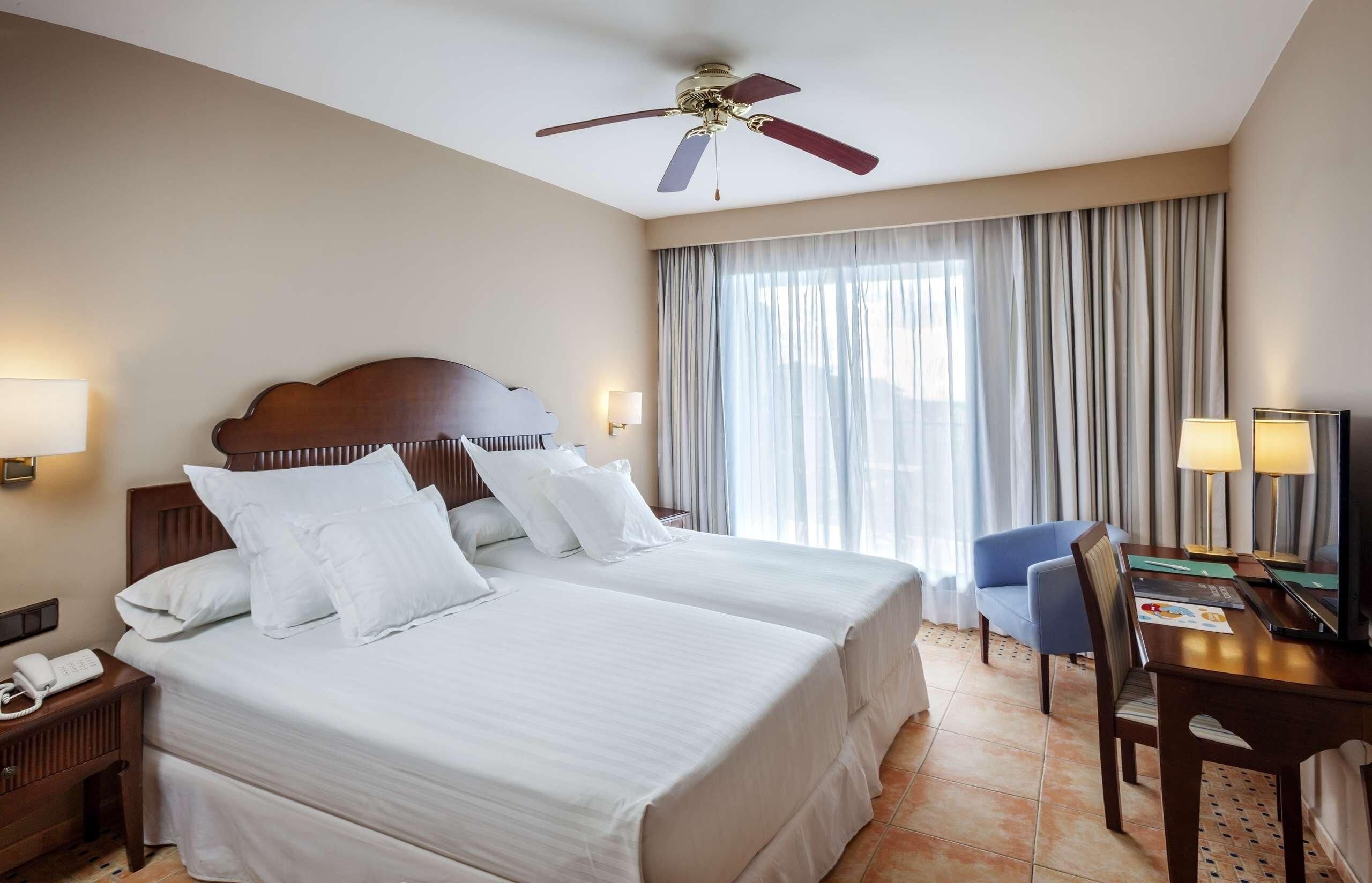 A hotel room with two beds, a desk, a chair, a ceiling fan, and a large window with curtains.