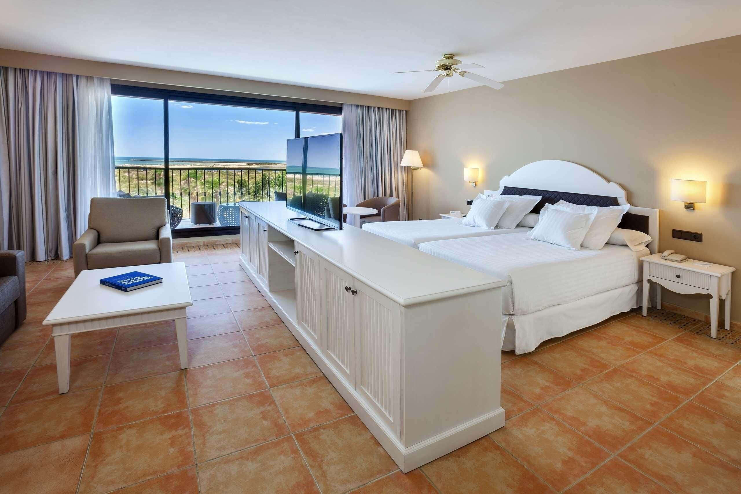 Habitación de hotel con cama, televisión, escritorio, silla y balcón con vistas.