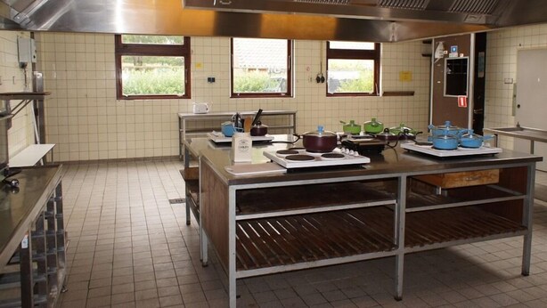 A commercial kitchen with stainless steel appliances, tiled walls, and windows with views of greenery. - The Budget Hotel (Hazerswoude-Rijndijk)