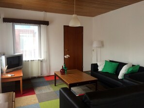 Ein Wohnzimmer mit Fernseher, einem Holztisch, einem schwarzen Sofa mit grünen und weißen Kissen, einer Holztür und einem Fenster mit Vorhängen.
