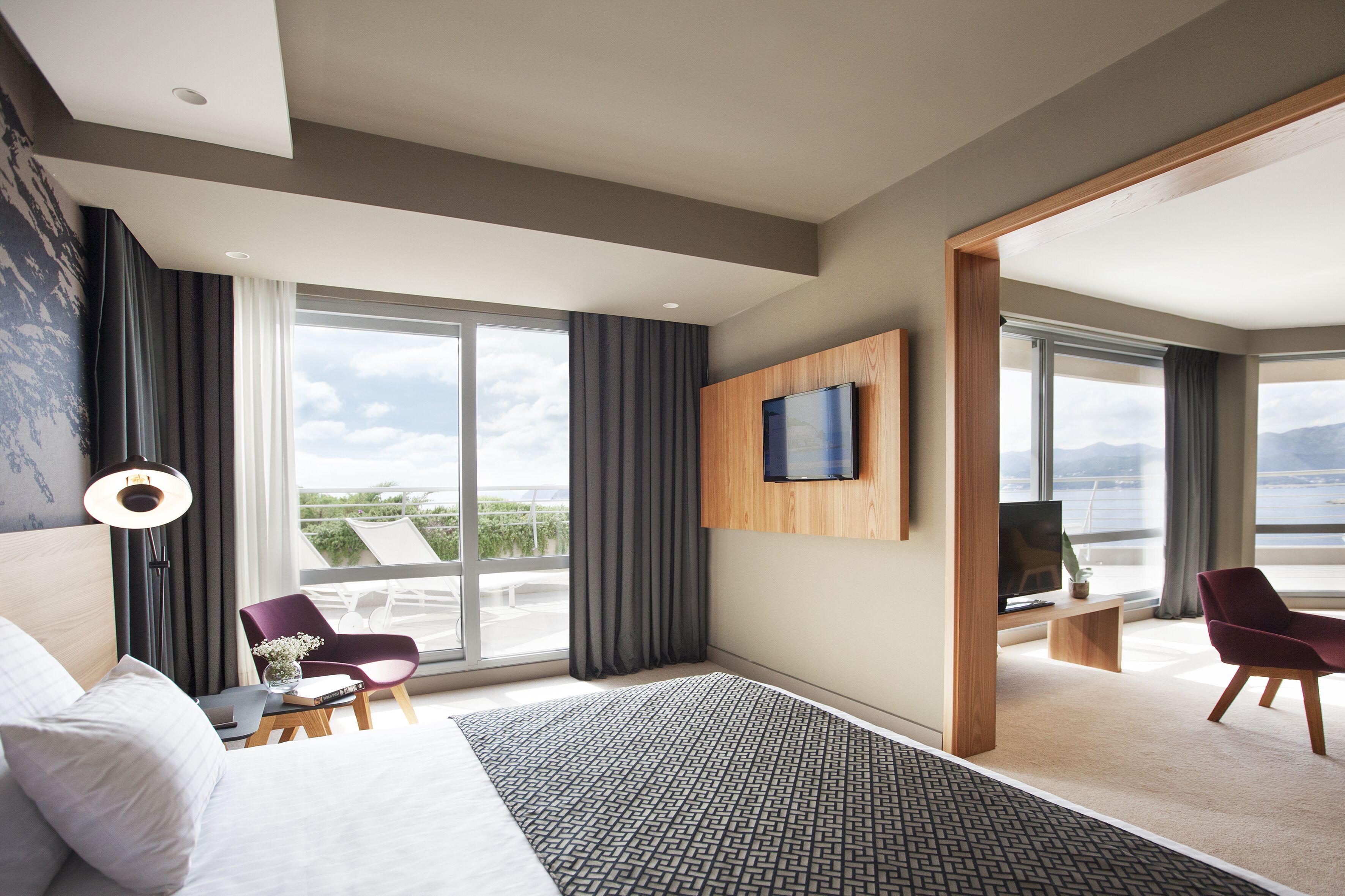 Quarto de hotel moderno com uma cama grande, uma mesa de trabalho, uma cadeira, TV fixada na parede e vista para o exterior.
