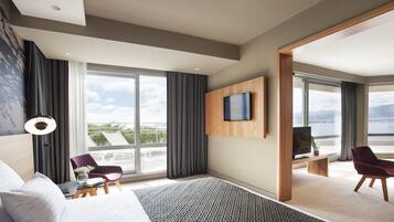 Quarto de hotel moderno com uma cama grande, uma mesa de trabalho, uma cadeira, TV fixada na parede e vista para o exterior.
