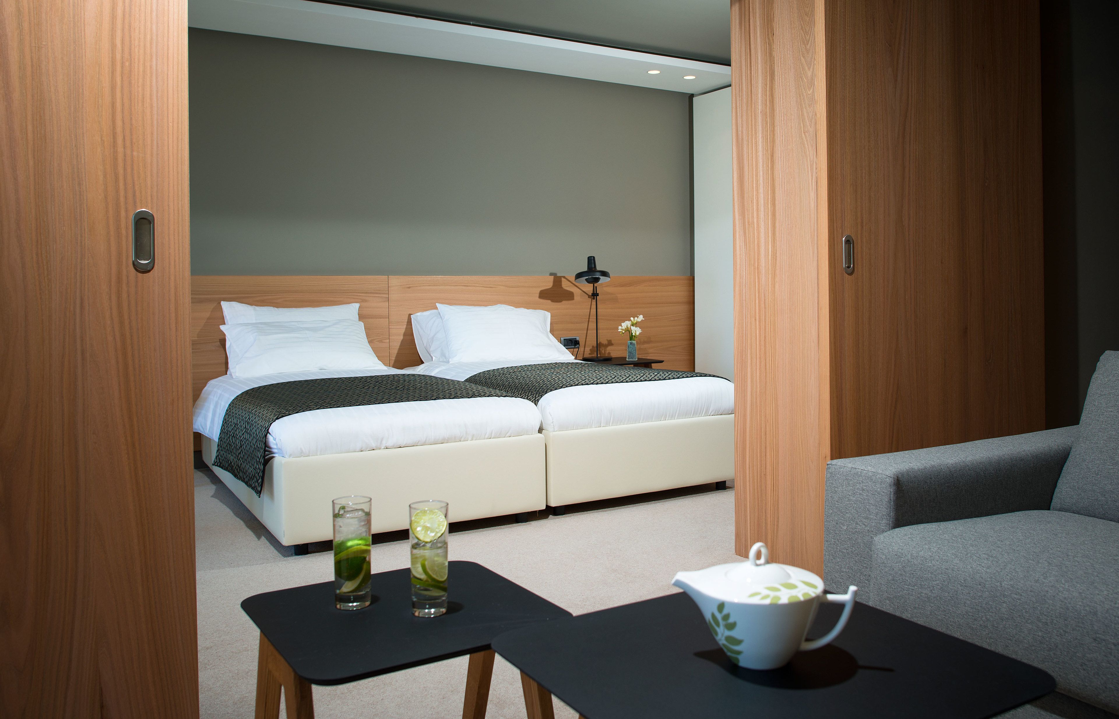 A modern hotel room with a double bed, a bedside table with a lamp, a small table with a teapot, and a chair.