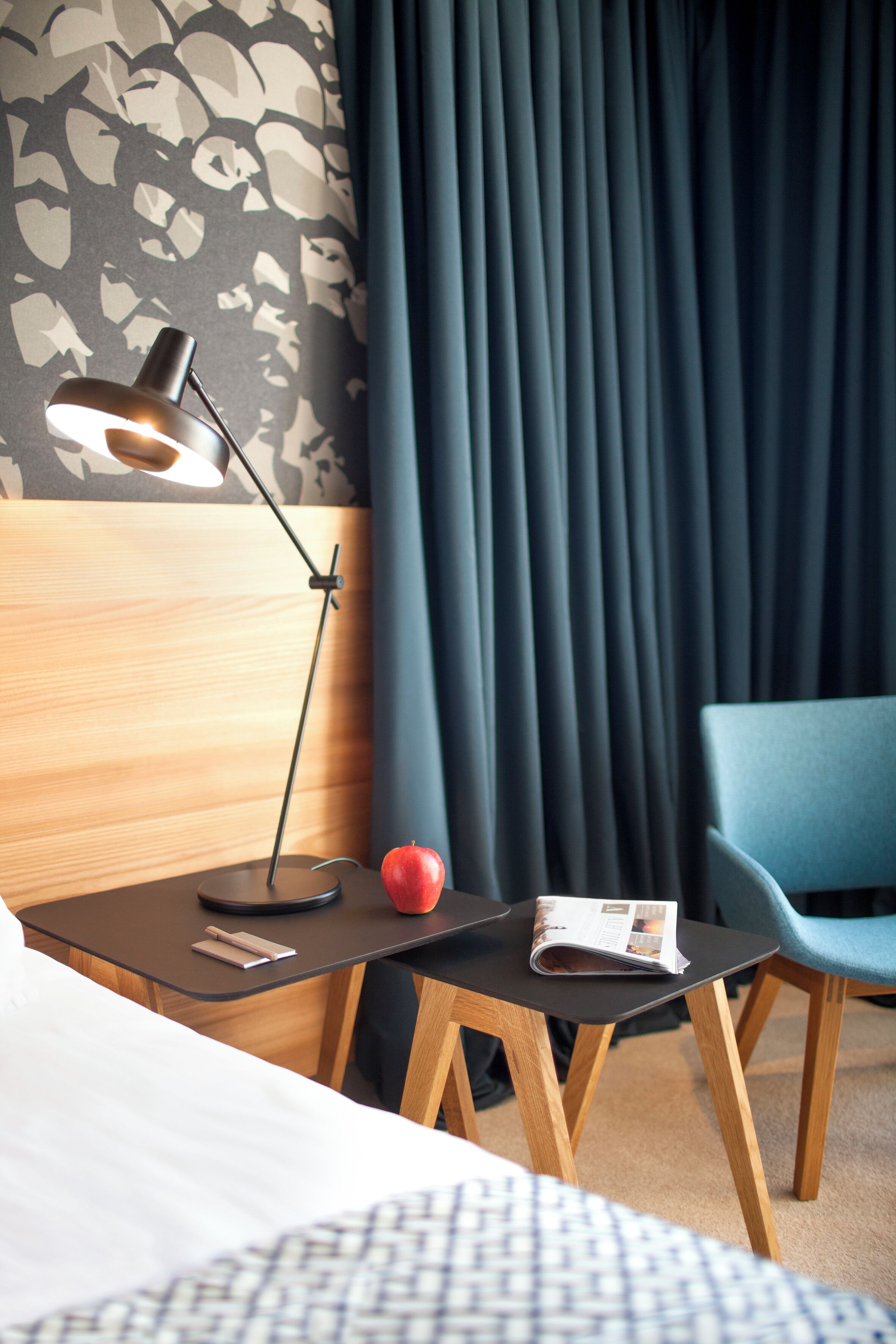 A hotel room with a bed, a desk with a lamp, a chair, a red apple, and a magazine.