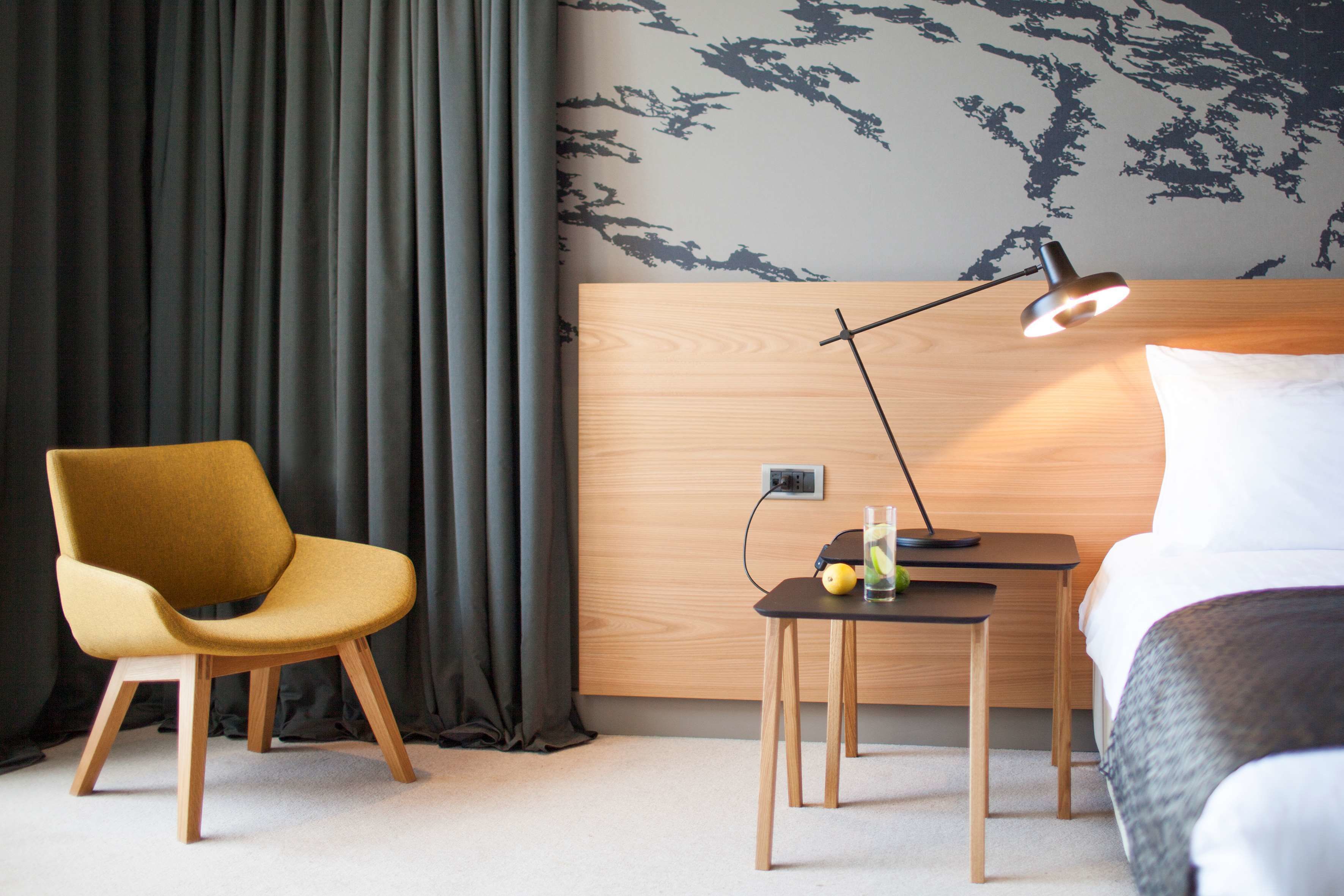 Ein Hotelzimmer mit Holz-Kopfteil, einem gelben Sessel, einem Nachttisch mit Lampe, einem Bett und einer Wand mit Blätermotiv.