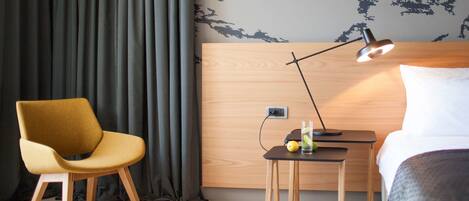 Ein Hotelzimmer mit Holz-Kopfteil, einem gelben Sessel, einem Nachttisch mit Lampe, einem Bett und einer Wand mit Blätermotiv.
