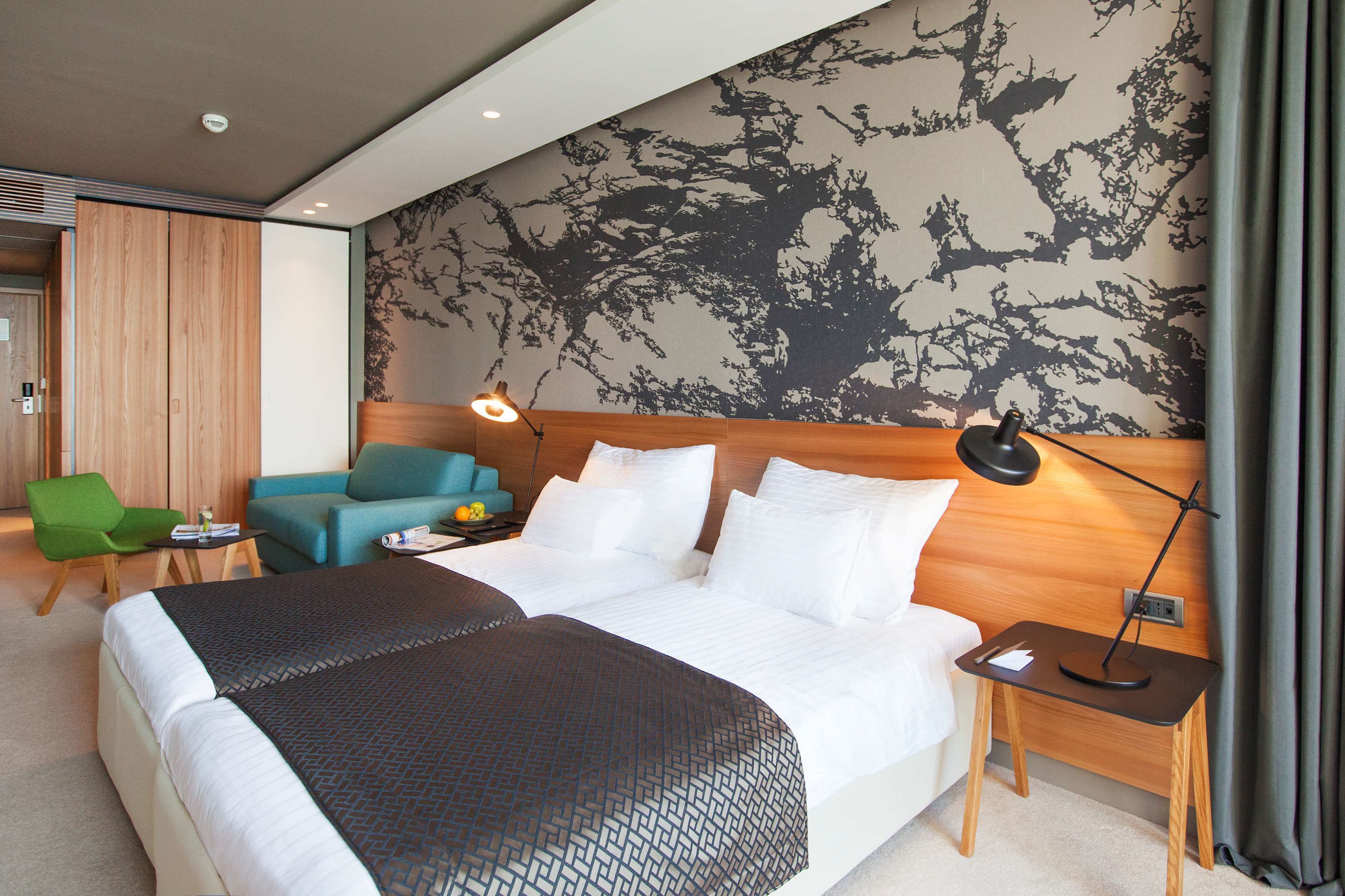 Une chambre d’hôtel avec un grand lit, une tête de lit en bois, une table de chevet avec une lampe, un fauteuil vert et un mur orné d’un motif représentant un arbre.