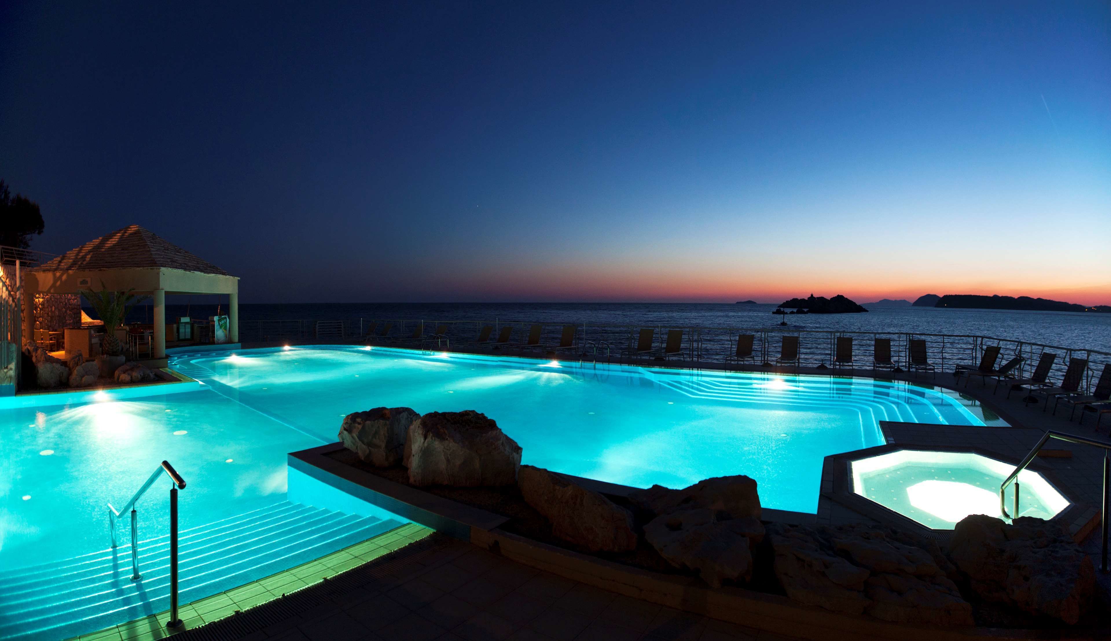 Piscina à noite com vista para o pôr do sol, cadeiras de descanso e um pequeno pavilhão.