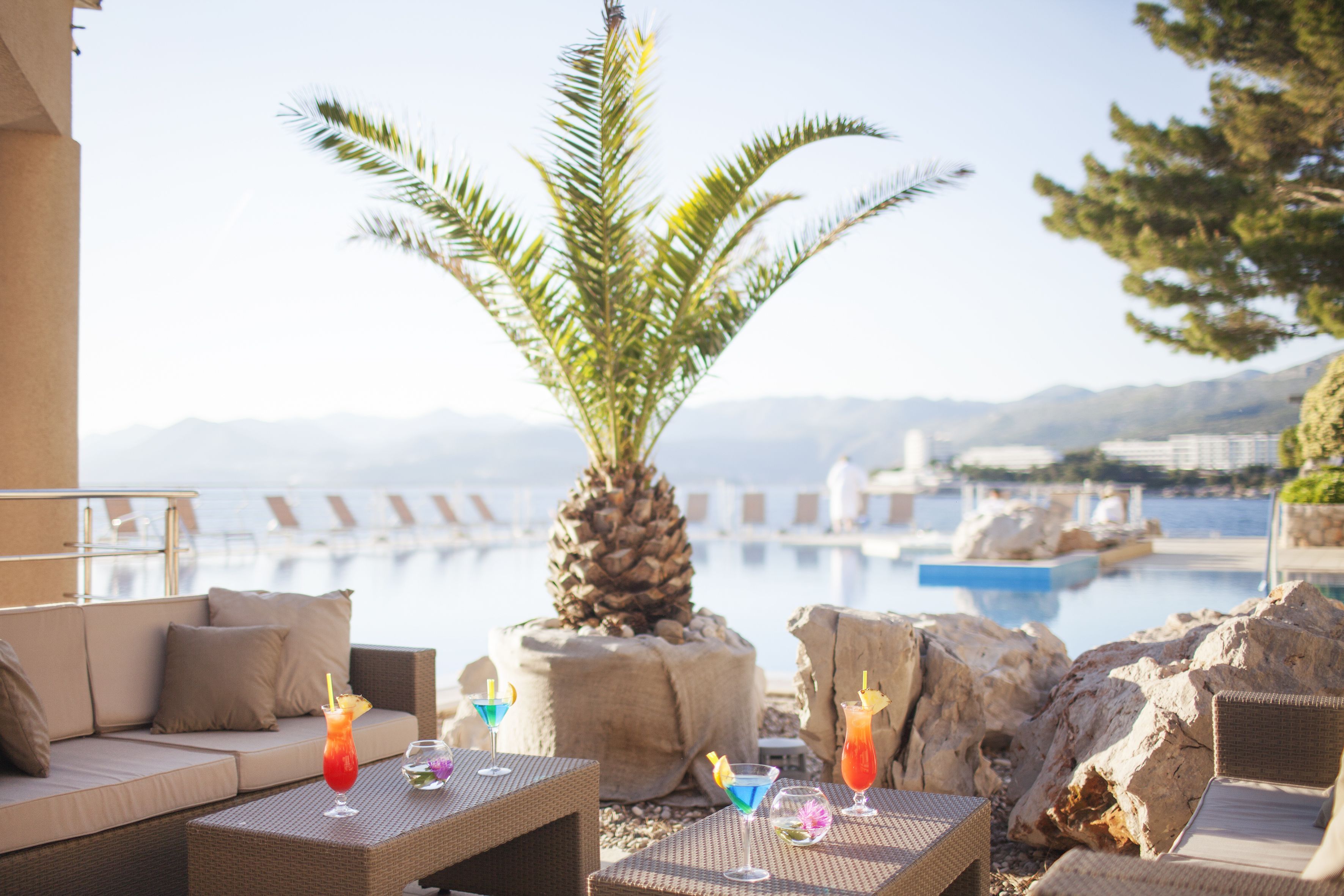 A poolside lounge area with wicker furniture, a palm tree, and a view of the pool and mountains in the background.
