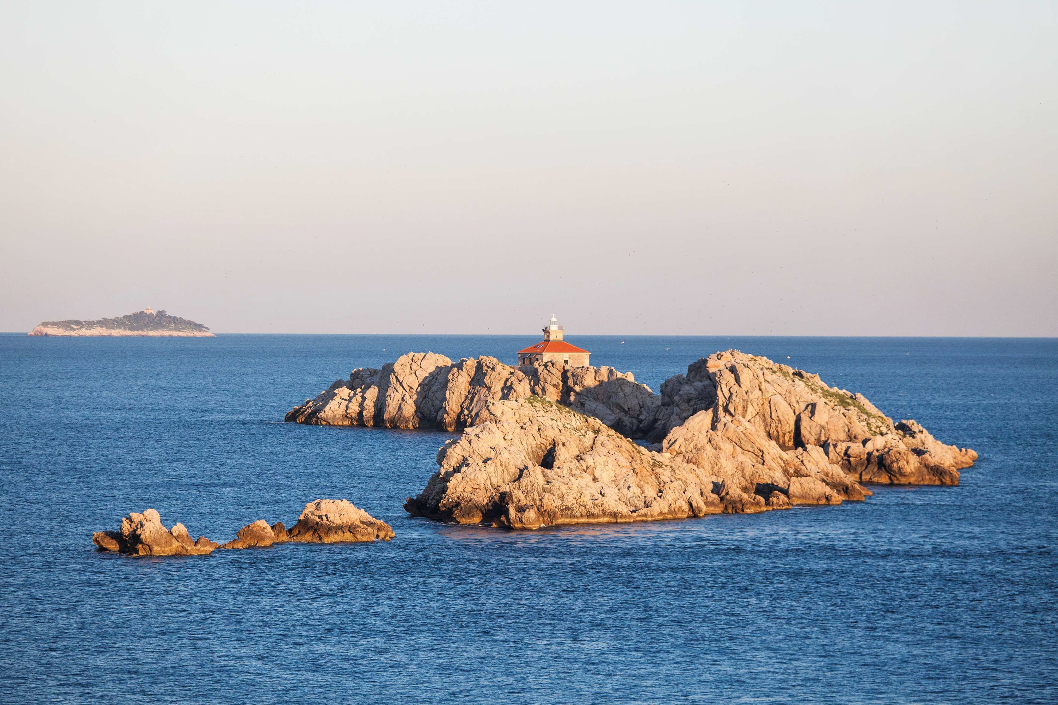 Uma ilha rochosa com um farol no centro, cercada por águas azuis calmas.