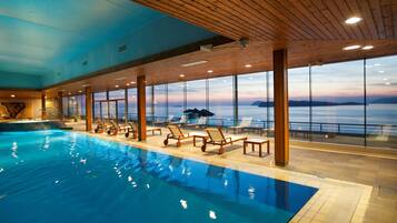 An indoor swimming pool with wooden deck chairs and large windows offering a view of the sea.