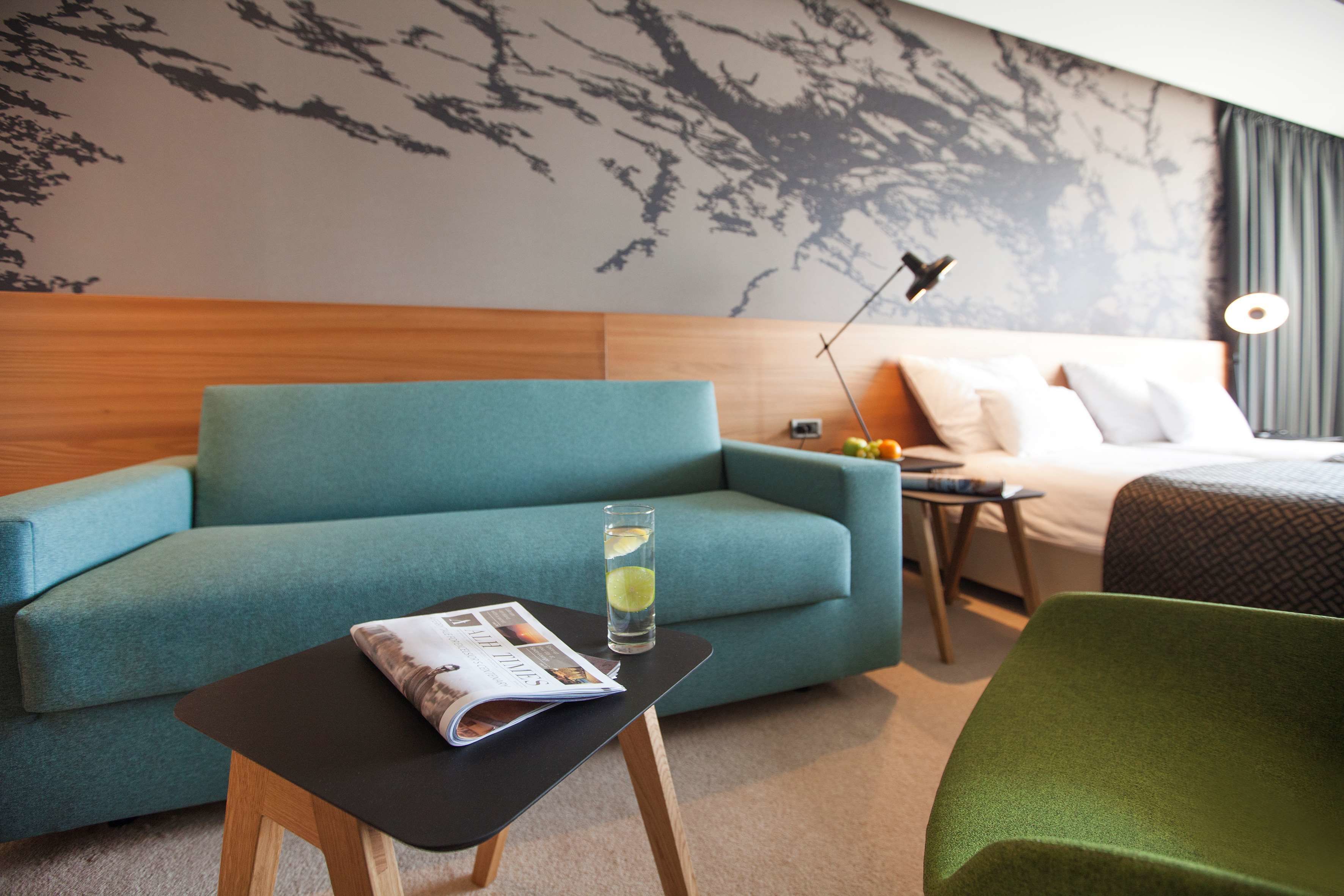 Una habitación de hotel moderna con un sofá azul turquesa, una mesita con una revista, un vaso de agua y una silla verde.
