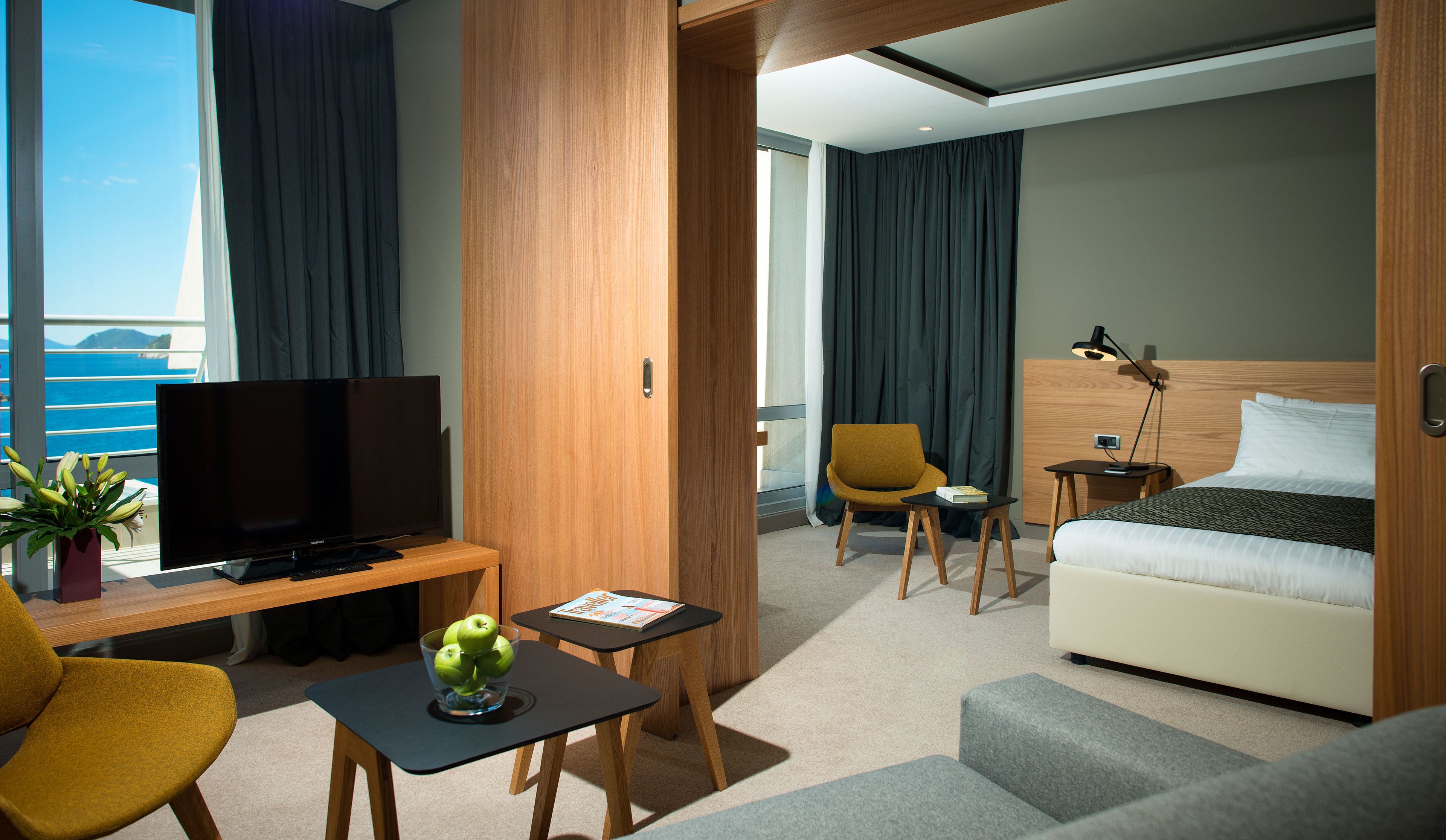 Ein modernes Hotelzimmer mit Fernseher, Sofa, Sesseln und einem kleinen Tisch mit einer Zeitschrift.