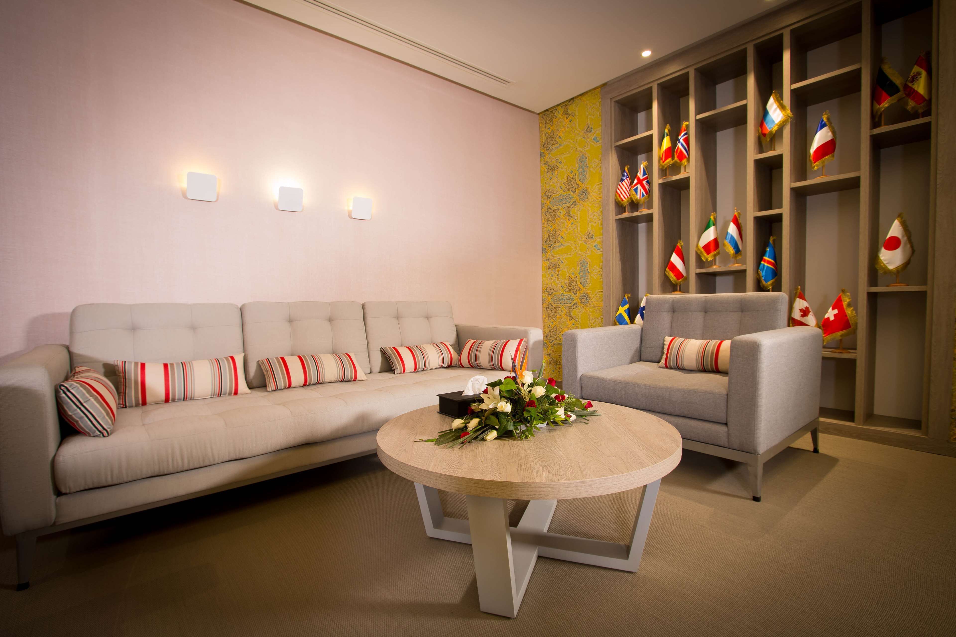 A modern living room with a sofa, armchair, and a round coffee table. There are wall-mounted lights and a shelf with flags.
