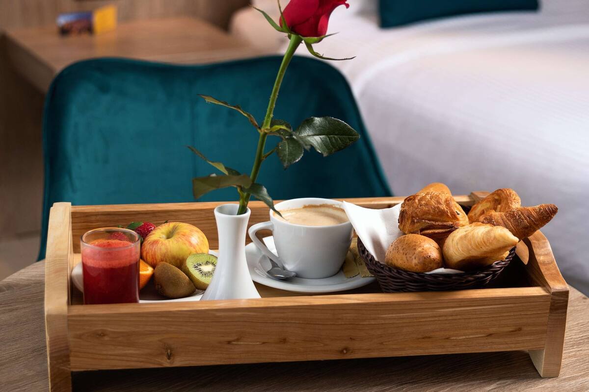 A breakfast tray with croissants, a cup of coffee, a red rose, and a fruit bowl on a wooden table in a hotel room.