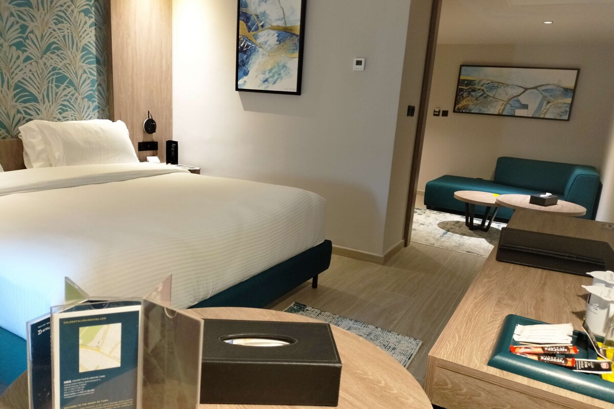A hotel room with a bed, a desk, a chair, a sofa, and artwork on the walls.