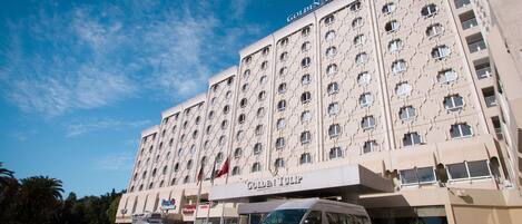 A large building with the sign "Golden Tulip" indicating it is a hotel. There are multiple cars parked in front, and a van is parked in front of the entrance.