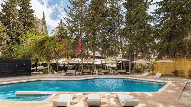 Una piscina con sillas de descanso y sombrillas, rodeada de árboles y un edificio.