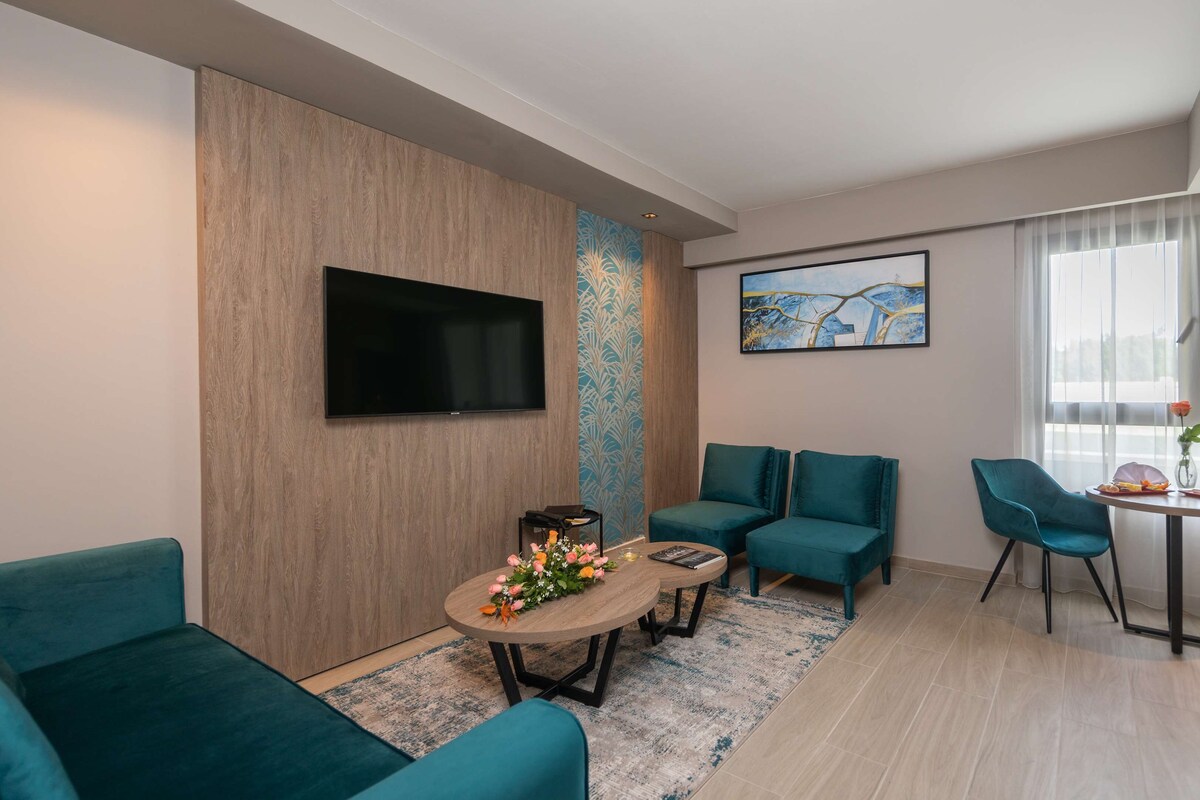 A modern living room with teal sofas, a coffee table with flowers, a flat-screen TV, and a painting on the wall.