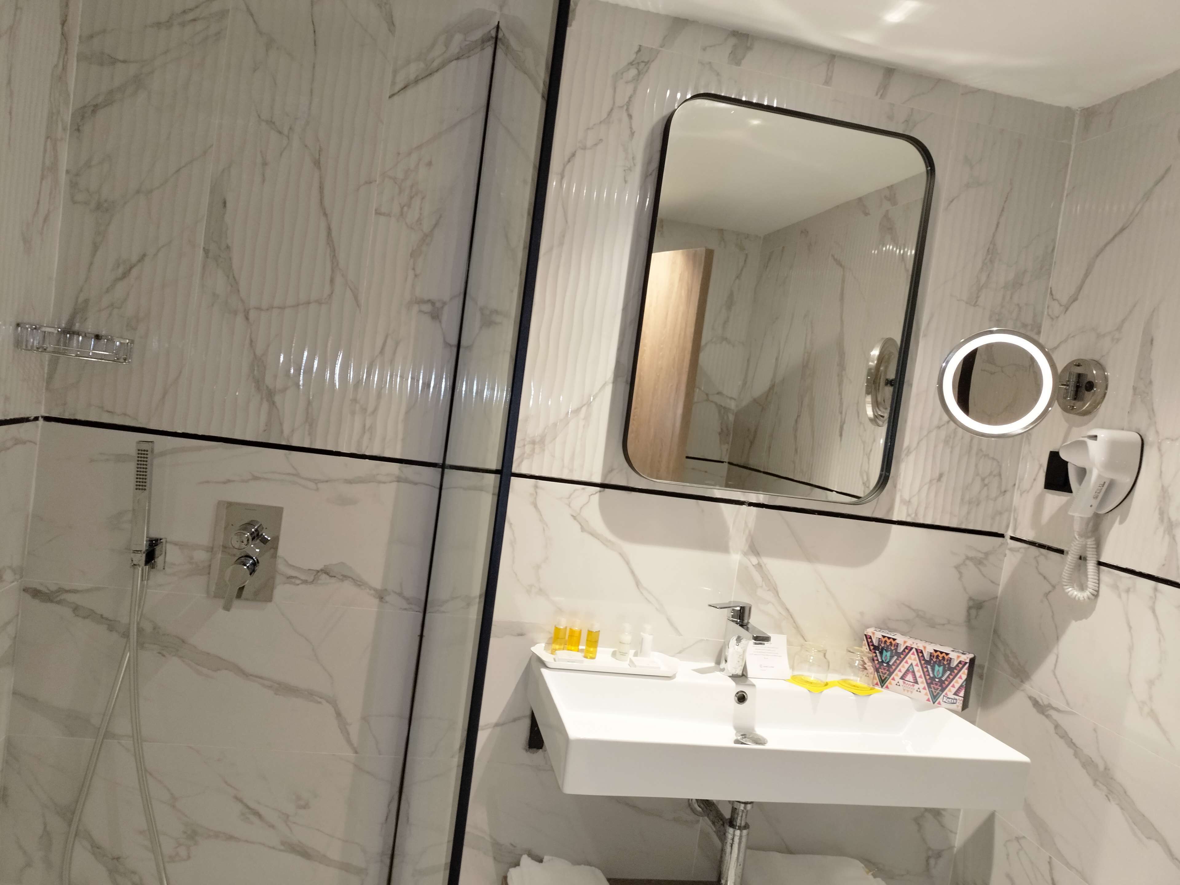 A bathroom with marble walls, a white sink, a handheld showerhead, a mirror, and a hairdryer.