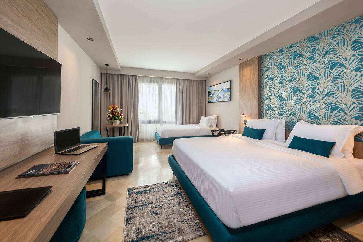 A hotel room with a large bed, a desk with a laptop, a TV, and a small table with a flower arrangement.