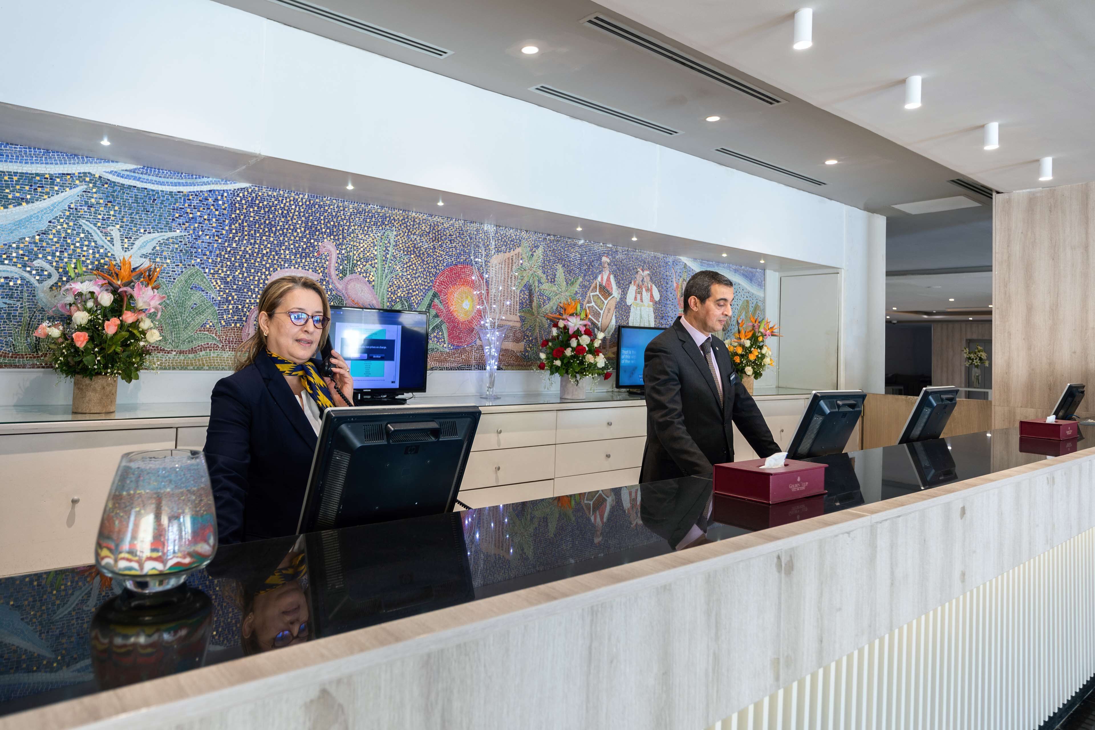 A reception desk with two monitors, a glass-top counter, and a mosaic wall in the background.
