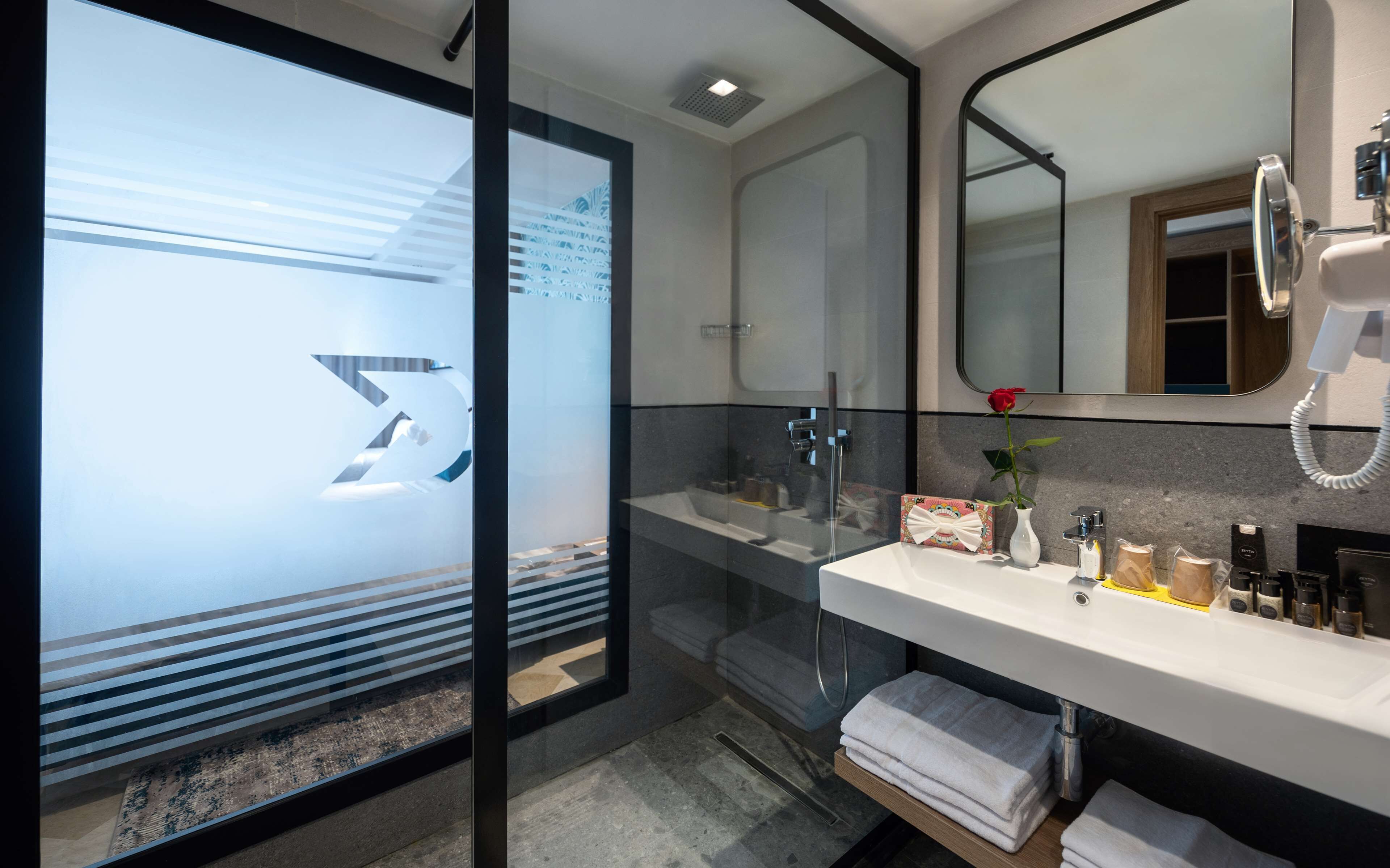 A modern bathroom with a glass shower enclosure, a vanity with a mirror, and a rose in a vase.