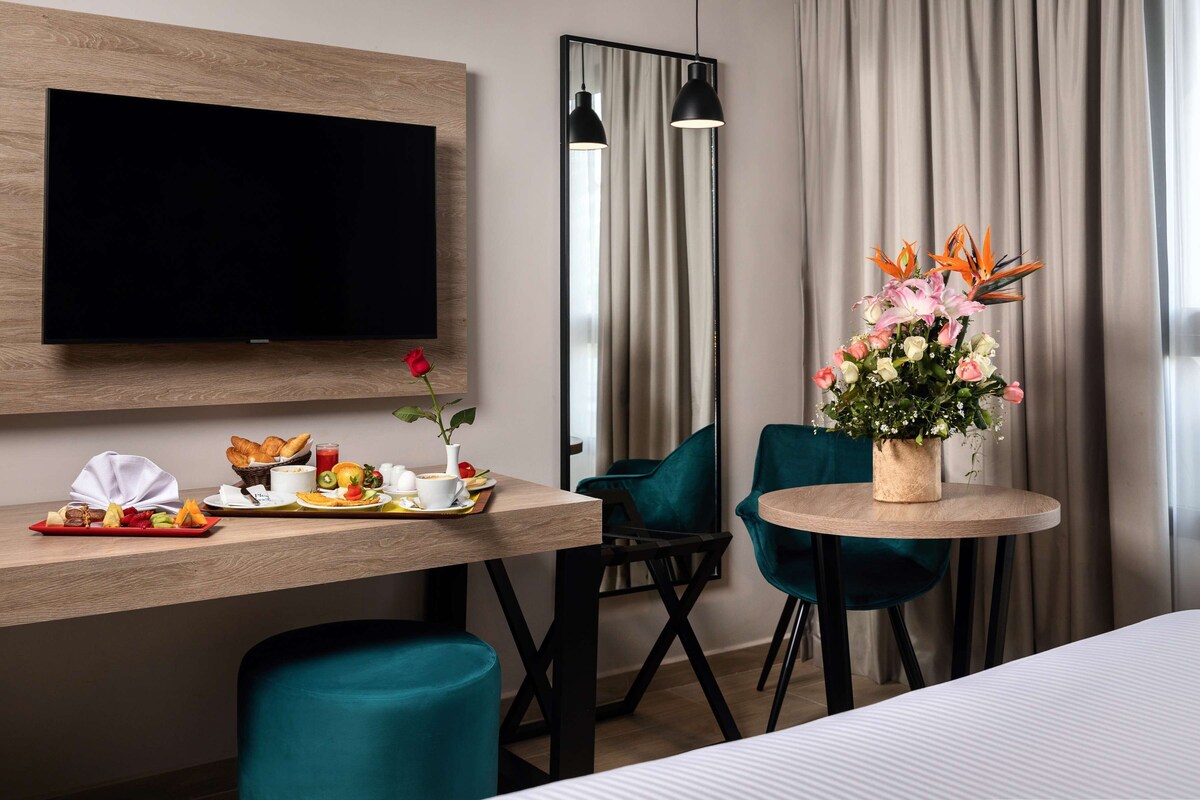 A hotel room with a flat-screen TV mounted on the wall, a dining table with breakfast items, teal chairs, and a flower vase.