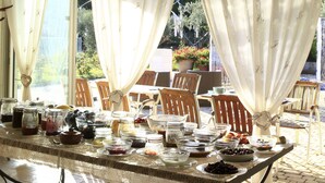 Un espacio exterior cubierto para comer con una mesa lista para una comida, rodeada de sillas y grandes ventanales con cortinas.