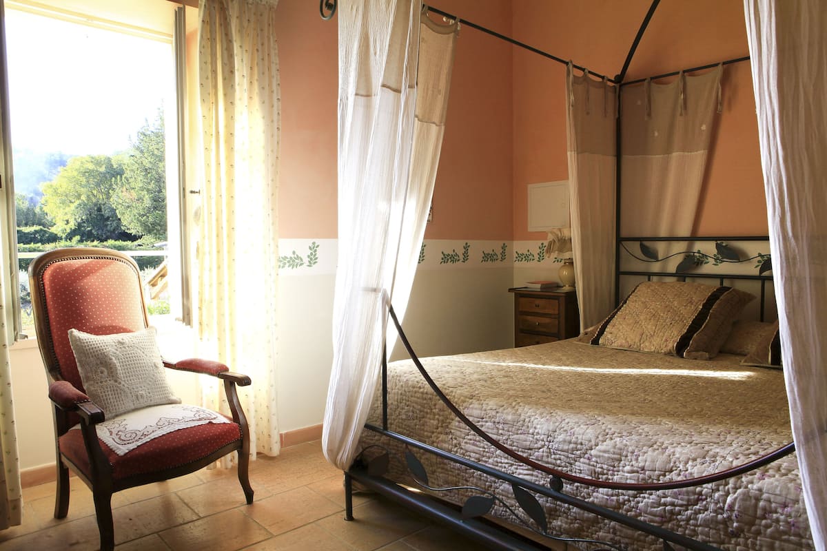 Ein Schlafzimmer mit einem Himmelbett, einem Holzstuhl mit Kissen und Blick ins Grüne.