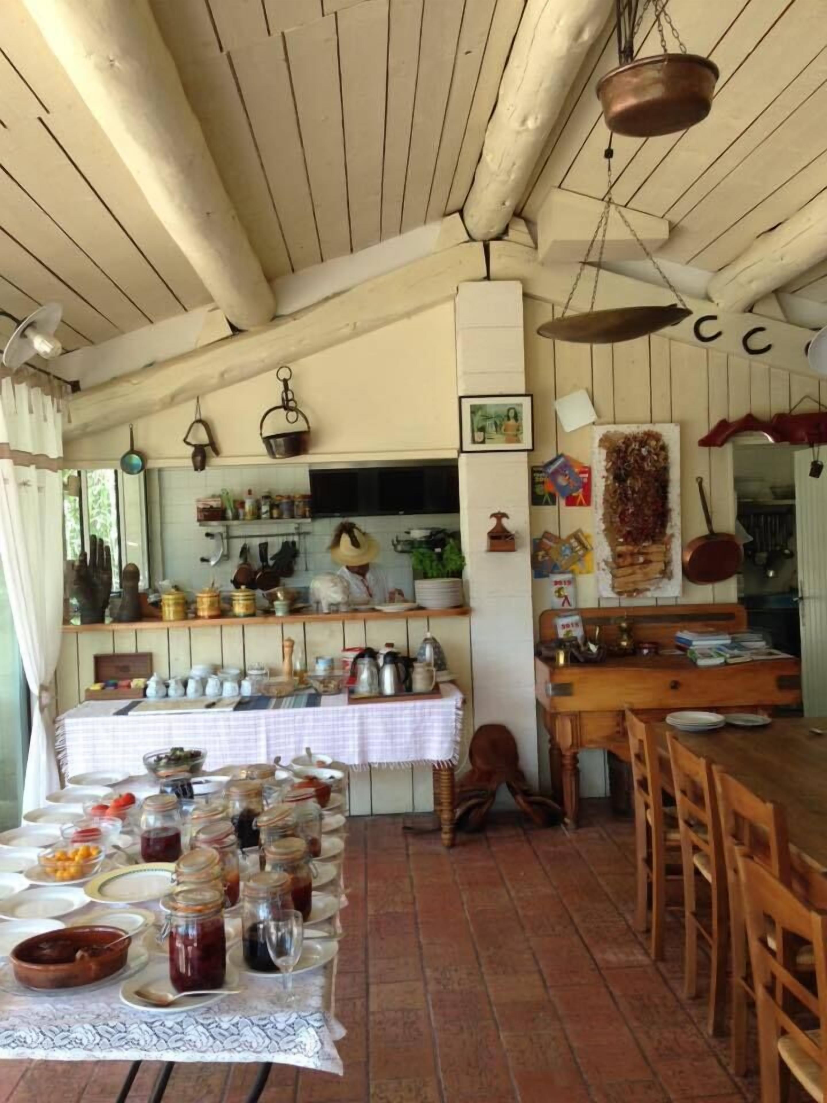 Une cuisine rustique avec des poutres en bois, une longue table dressée pour le petit-déjeuner et divers ustensiles de cuisine suspendus au-dessus.