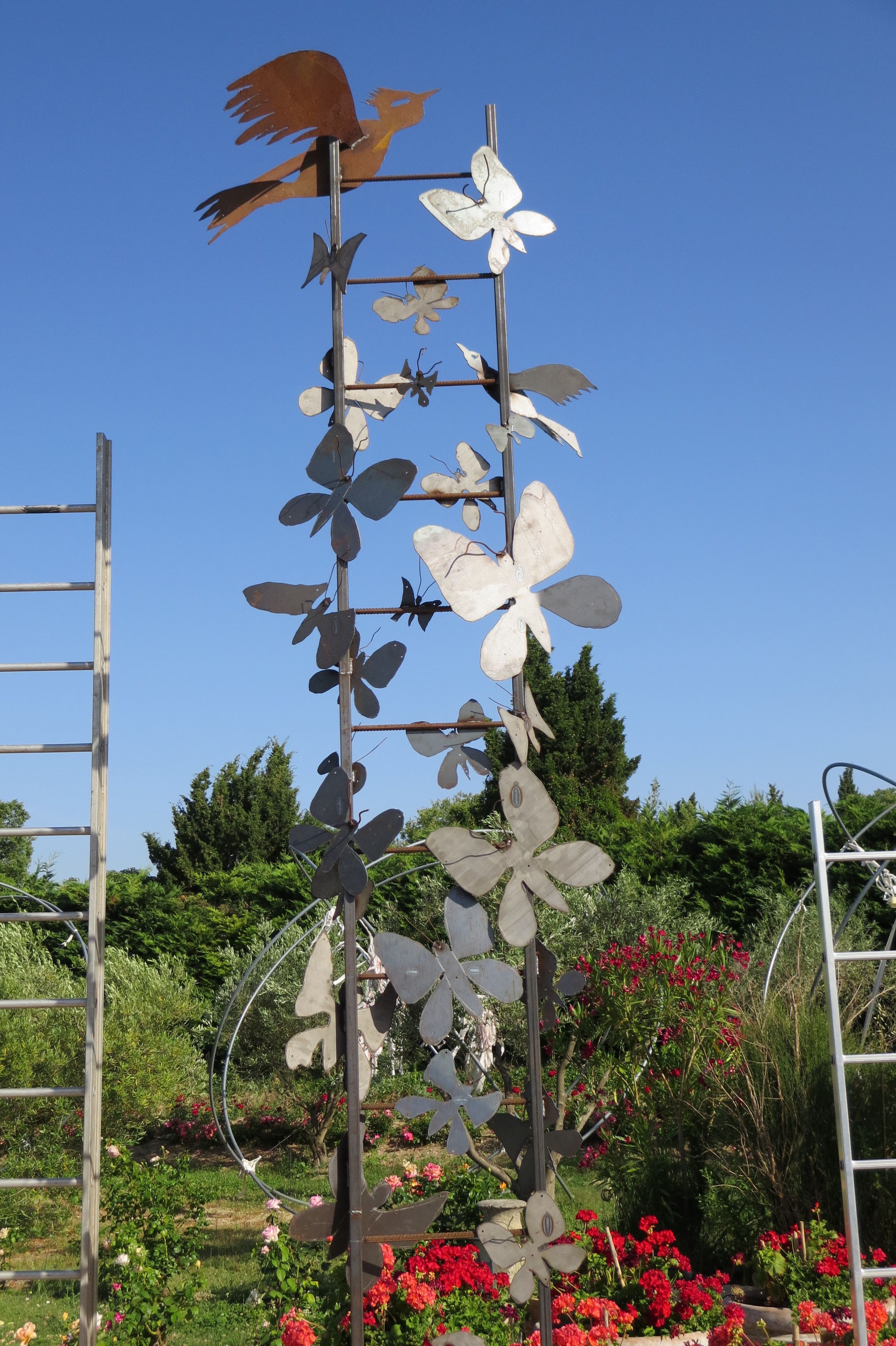 Escultura de metal com pássaros e flores num jardim.