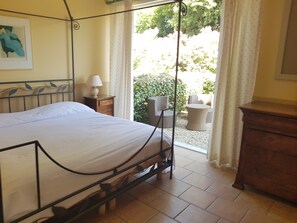 A bedroom with a bed, a nightstand with a lamp, a dresser, and a view of an outdoor patio with wicker furniture and greenery.